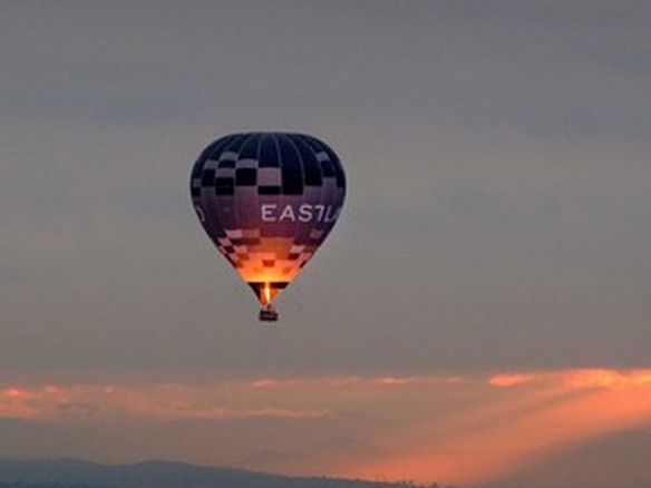 A hot air balloon floats above a mountainous landscape at sunset, with warm rays of sunlight breaking through the clouds.