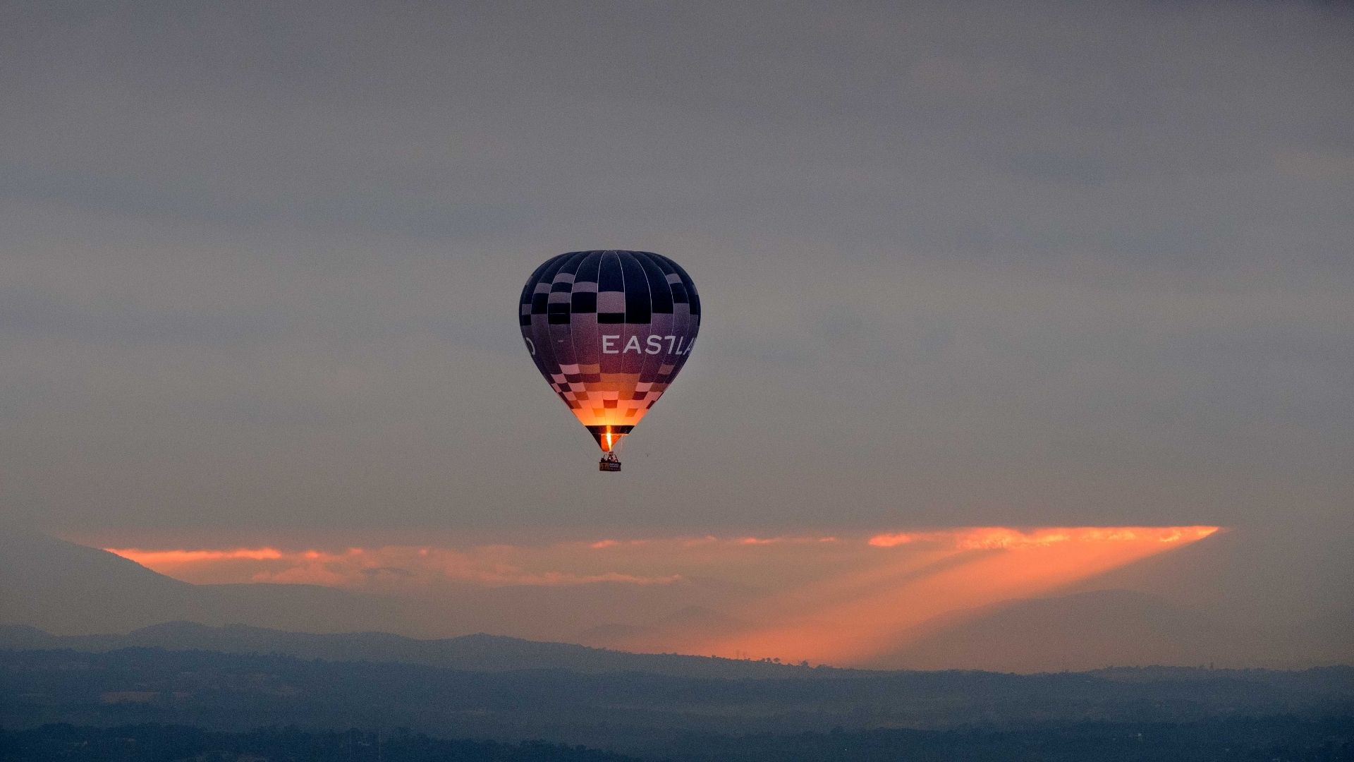 A hot air balloon floats above a mountainous landscape at sunset, with warm rays of sunlight breaking through the clouds.