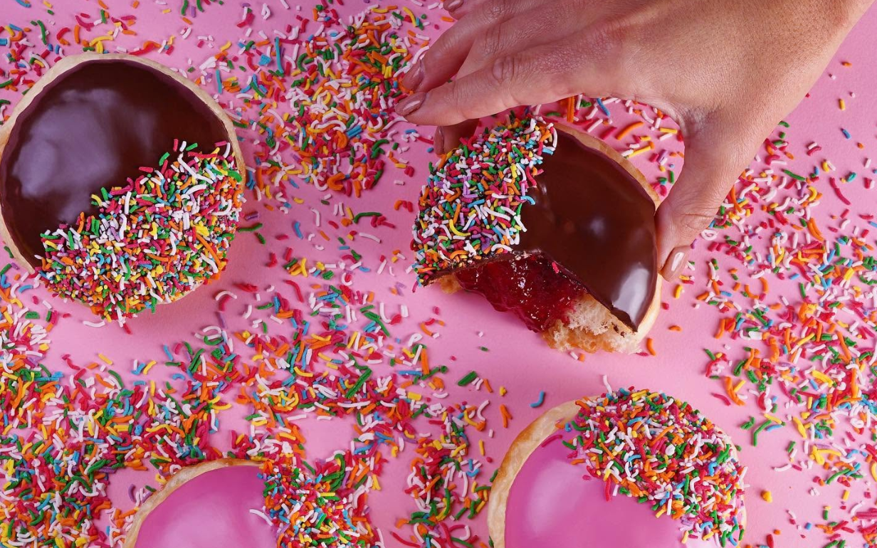 Colourful iced donuts covered in rainbow sprinkles, with a hand decorating one donut.