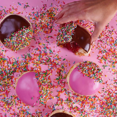 Colourful iced donuts covered in rainbow sprinkles, with a hand decorating one donut.