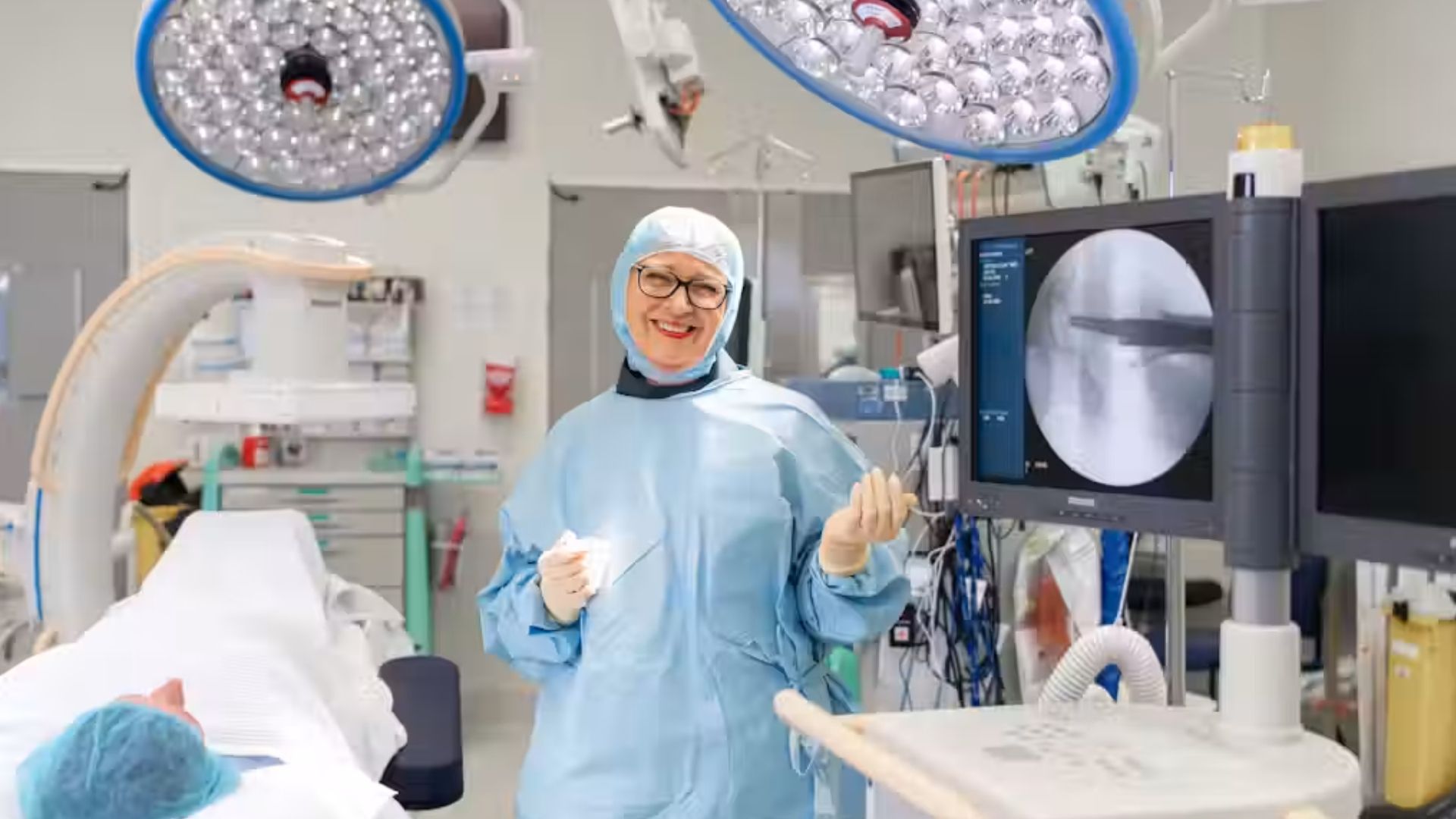 A smiling healthcare professional in surgical scrubs stands in a hospital operating room beside medical imaging equipment.