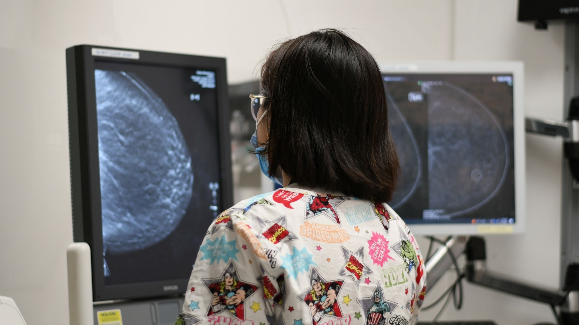 A healthcare worker views mammogram images displayed on two computer monitors in a clinical setting.