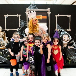 A group of children in colourful Halloween costumes pose with a costumed character in front of a spooky Halloween display decorated with skeletons, spider webs and bones inside a shopping centre.