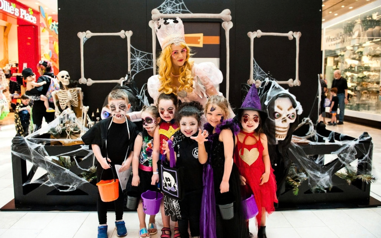 A group of children in colourful Halloween costumes pose with a costumed character in front of a spooky Halloween display decorated with skeletons, spider webs and bones inside a shopping centre.