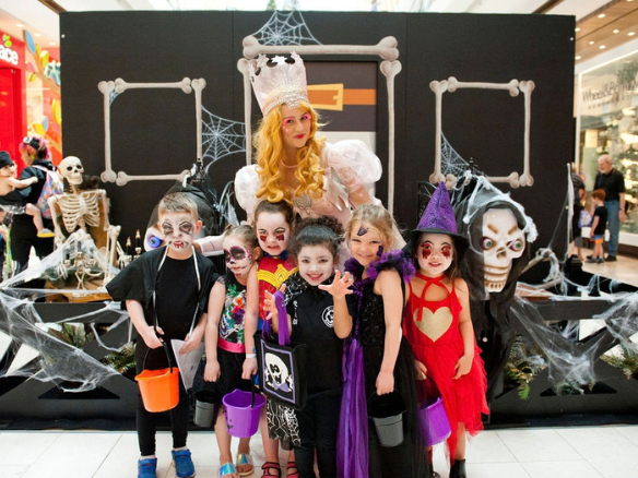 A group of children in colourful Halloween costumes pose with a costumed character in front of a spooky Halloween display decorated with skeletons, spider webs and bones inside a shopping centre.