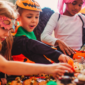 Three children in Halloween costumes gather around a table filled with colorful treats, reaching for food and candy.