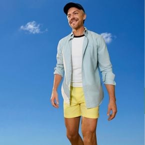 Smiling man wearing a navy cap, pale blue button-up shirt and white T-shirt, pictured against a bright blue sky with clouds.