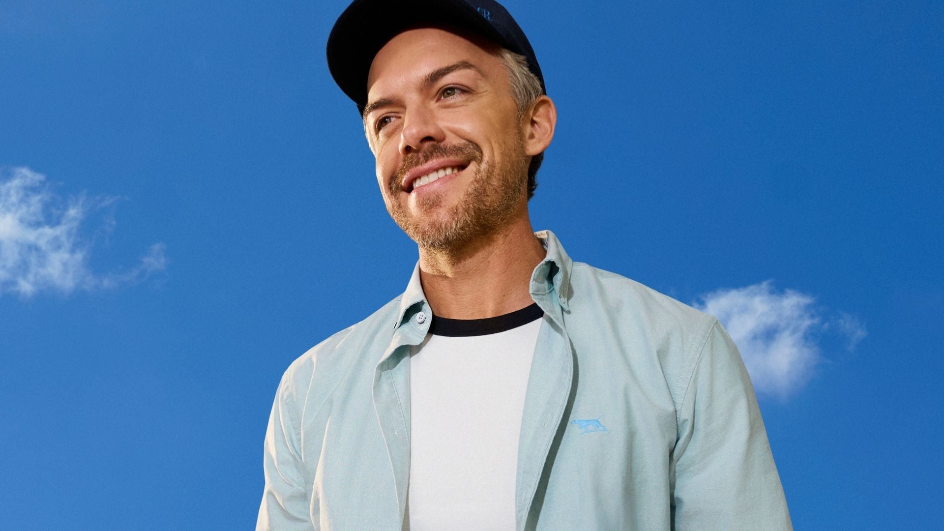 Smiling man wearing a navy cap, pale blue button-up shirt and white T-shirt, pictured against a bright blue sky with clouds.