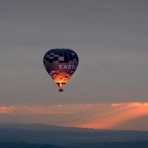 A hot air balloon floats above a mountainous landscape at sunset, with warm rays of sunlight breaking through the clouds.