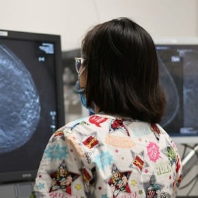 A healthcare worker views mammogram images displayed on two computer monitors in a clinical setting.
