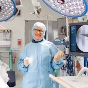 A smiling healthcare professional in surgical scrubs stands in a hospital operating room beside medical imaging equipment.